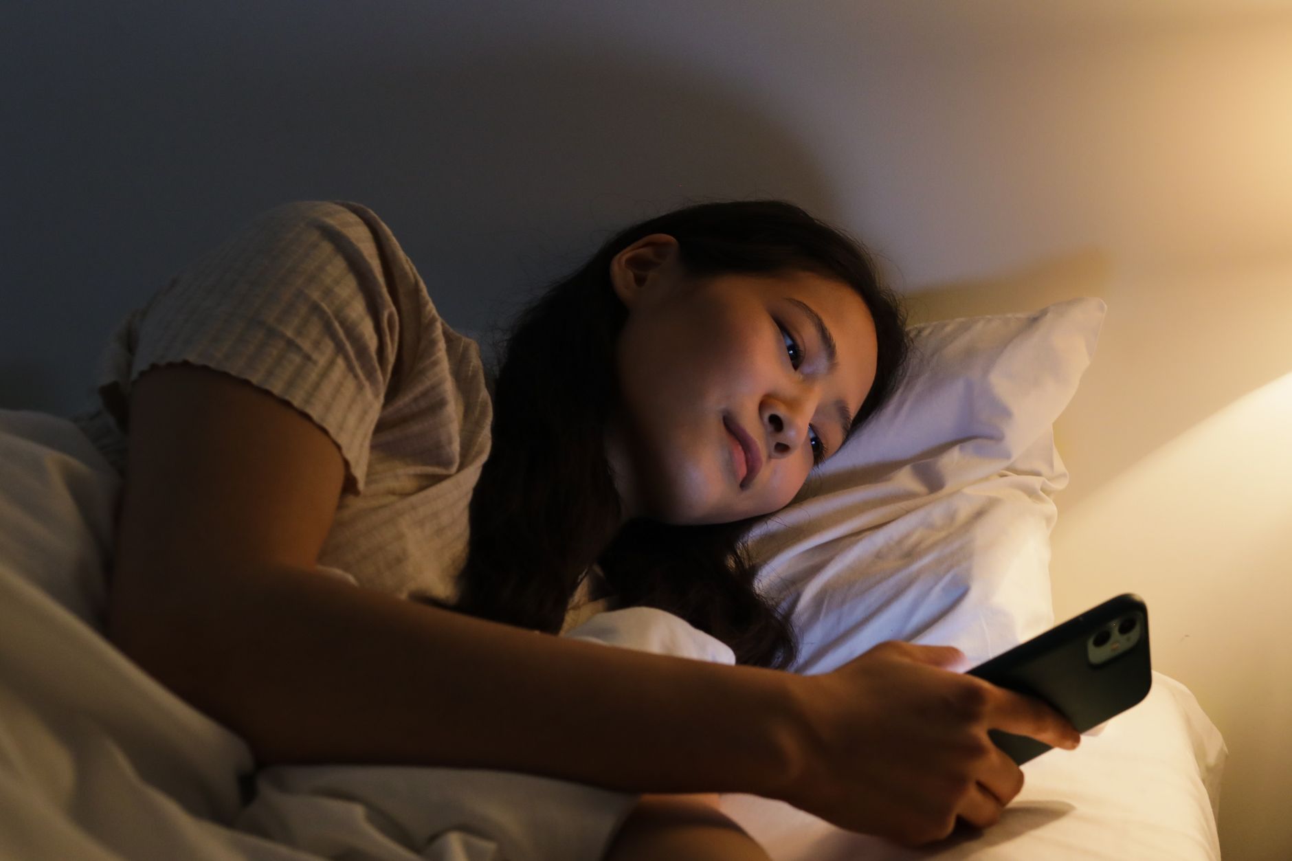 暗い部屋でベッドに入ったままスマートフォンを見ている女性のイメージ
